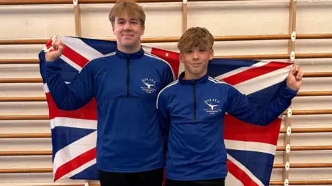 Splitz Gymnastics Club Archie and Samuel in Splitz Gymnastics Club tracksuits standing together holding a Union Jack draped behind them in a gymnasium