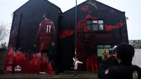 Reuters A man takes a photo on his mobile phone of a woman imitating Salah's celebration pose of arms spread out, which s depicted on a huge mural that covers the end of a household terrace. The red and black mural shows Salah wearing a golden crown and another image of him from the back with his LFC shirt and his name and number 11 on the back.
