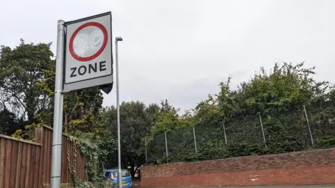 Edward Lamb A 20mph speed limit sign on a suburban road is daubed with white paint, rendering the number 20 harder to see. There is a wall, fencing and trees in the background