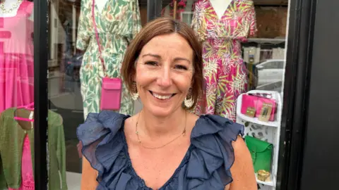 Ben Parker/BBC A woman with a blue top is standing in front of a shop window with mannequins wearing bright clothing.