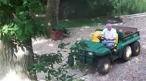 John Palmer in a white t-shirt is driving a green ride-on lawnmower on the gravel of his garden on a sunny day. A time code on the picture reads: 24-06-2015. Wed. 16:23:42.