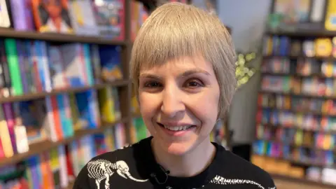 Author Thana Niveau with short grey hair smiling at the camera wearing a dark top with Skeleton animals on it