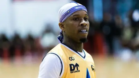 A man wears a yellow basketball jersey over a white t-shirt, and a blue-and-white bandana covering his head. A section of basketball court and a row of spectators can just be made out in the background.