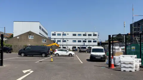 An image of Oasis Academy Temple Quarter, which is a white building with square windows. The view is from the car park where you can see a few cars and vans parked.