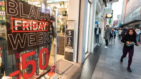 Getty Images A woman walks past a sign on the high street that says Black Friday Week - 50% and the work Friday is crossed out