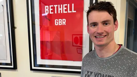 Dan Bethell has short dark hair and is smiling at the camera. He is standing beside a framed red t-shirt that says "Bethell" and is signed by him.