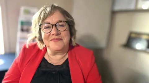 Martin Giles/BBC A woman sitting in an office. She is wearing a red jacket and a black top. She is wearing glasses and smiling.