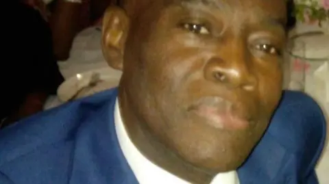 The image shows a black man wearing a blue suit with a white shirt and multi-coloured tie.