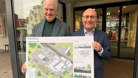 Cllr John Gallagher and Fred Moroni, Chief Executive Officer at Malvern Theatres stand outside the theatre with a plan of the redevelopment. The glass doors of the theatres are behind them and there are lights on inside.
