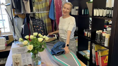 A woman stands smiling behind an ironing board. There are racks of clothes behind her and a vase of white roses to one side. She is holding an iron in one hand and a pair of jeans with the other.