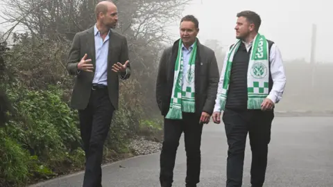 PA Media Prince William is talking as he walks down the new access road with two men from the football club who are wearing the team's scarves around their necks. It is foggy.