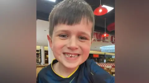 Archie smiles at the camera. He has short dark hair and a big smile on his face, showing a tooth missing from the bottom row. He is wearing a navy football shirt with yellow trim and appears to be in a restaurant