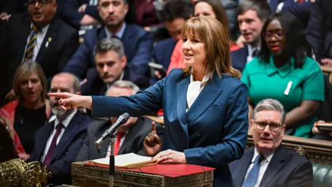 House of Commons/PA Wire Chancellor Rachel Reeves delivers the Budget in the House of Commons on 26 November 2025. She wears a blue suit and is pointing with her right hand, index finger extended. Prime Minister Sir Keir Starmer sits behind her on the front  bench, with other Labour MPs visible, listening to Reeves' speech. 
