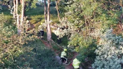 Cheddar Fire Station Five fire fighters wearing their hi-vis uniforms and helmets holding a long red rope. They are surrounded by trees and bushes