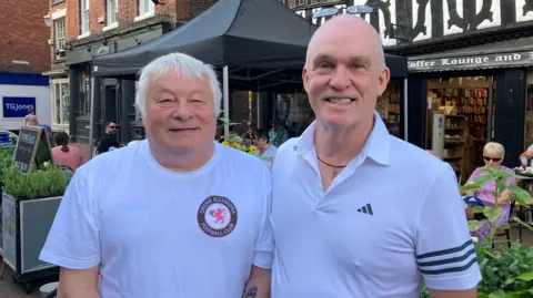 Two men in white t-shirts, stand next to each other outside a book shop, and are smiling. One has a Crewe Alex FC logo on his top.