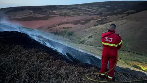 Derbyshire Fire and Rescue Service Firefighter with a hose standing next to smouldering moorland
