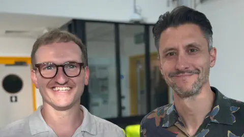 George King/BBC A head and shoulders image of two men - one wearing a white shirt and one wearing a dark coloured shirt with flower patterns on it. They are both smiling and looking into the camera.