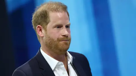 File photo of Prince Harry on a blue background, wearing a white open-neck shirt and a dark blazer.