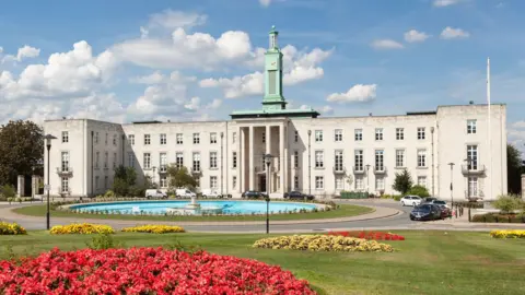 Getty Images A wide view of Waltham Forest town hall, large pale stone civic building with a central columned entrance and a green clock tower rising above it, seen across a circular fountain and manicured gardens with red and yellow flowerbeds under a blue sky.