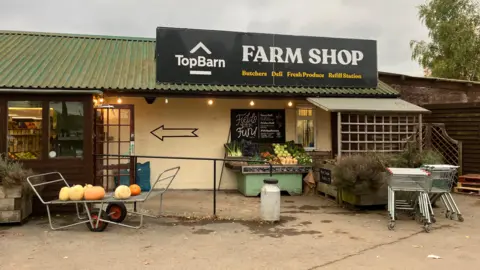 A low building with a sloping corrugated roof. A sign on top reads: "Top Barn Farm Shop - Butchers - Deli - Fresh Produce - Refill Station"