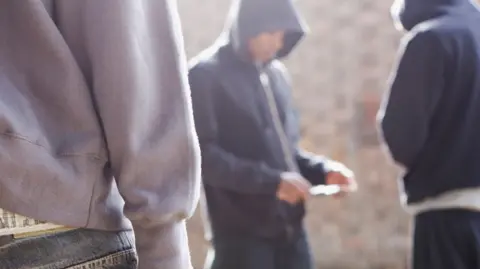 A blurred generic image of three young people wearing hooded tops. One of them appears to have a mobile phone in his hand.