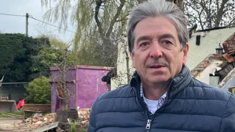 Tony Fisher/BBC A man with grey hair who is wearing a blue coat and a white checkered shirt. He is standing in front of a building which is in the process of being demolished and only has some walls remaining. There is a tree in the background.