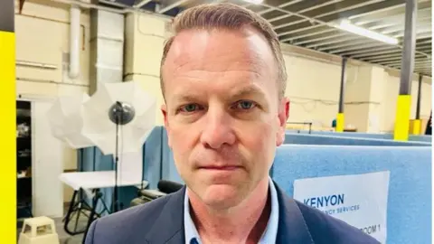 Linsey Smith/BBC Kevin Curreri stands in an industrial warehouse wearing a blue suit. Behind him are lights and a table.