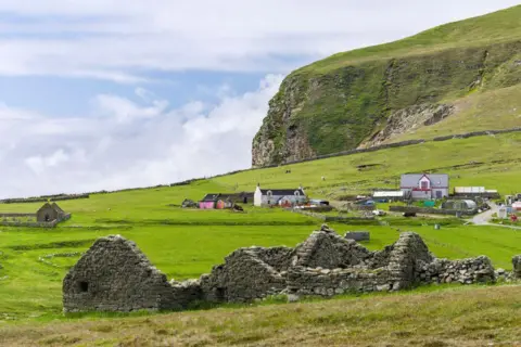 Getty Images Rumah pertanian terpencil di pulau Foula di Shetland, dengan reruntuhan bangunan pertanian di latar depan, di tengah ladang berumput, dan tebing tinggi berbatu di latar belakang.