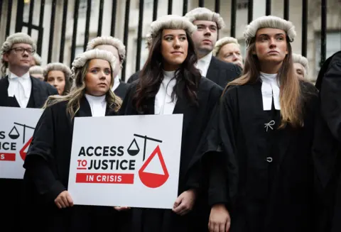 A row of barristers wearing black robes and wigs stand in front of a set of black iron railings. The three at the front are woman with long brown hair, behind them men can be seen. The woman in the middle is holding a placard which has a stylised scales of justice on it and reads "Access to Justice In Crisis"