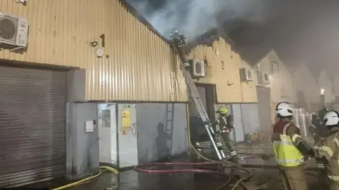 London Fire Brigade Firefighters respond to a fire at a warehouse - the roof is visibly damaged and smoke is rising from the building 