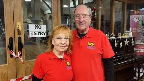 A man and woman stood arm in arm and both wearing red polo shirts with a logo on the right-hand side of their chests