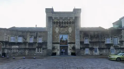 Google Maps A view of the front of Malmaison Oxford at Oxford Castle Quarter. A yellow car is parked to the side. It is a clear day.