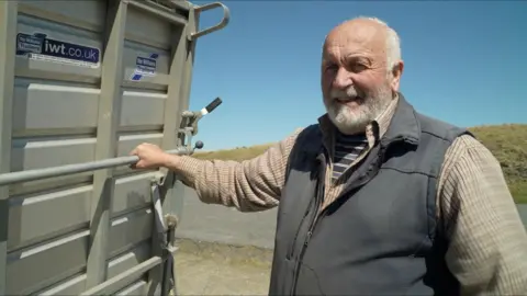 A man with a grey beard is pictured wearing a blue gilet and shirt. He is holding onto large machinery.