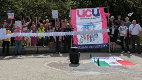 UCU members forming a picket line outside Durham University. Several people in the large group are carrying placards, while two people are supporting a large pink and white UCU banner.