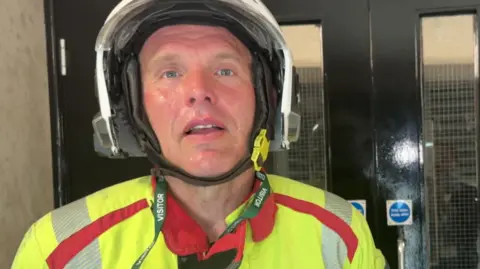 Fireman Samuel Davies speaks to the BBC while wearing a green jacket and white helmet