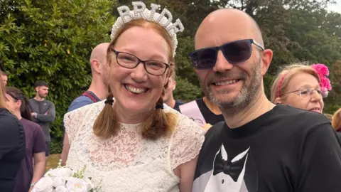 Sara Butler and Chris Hall are smiling. Butler is on the left and has ginger hair which has been tied into two plaits. She has dark glasses on and is wearing a white pearl headband which says 'BRIDE'. She is also wearing a lace cream dress with short sleeves and is holding a white bouquet of flowers. Hall is bald with a brown and grey short beard. He is wearing dark sunglasses and is wearing a black t-shirt which is designed to look like a tuxedo with a bow tie and white shirt. There are people standing around them. One woman behind Hall is blonde and is wearing a pink hairband with a large flower. There are trees and bushes behind them.