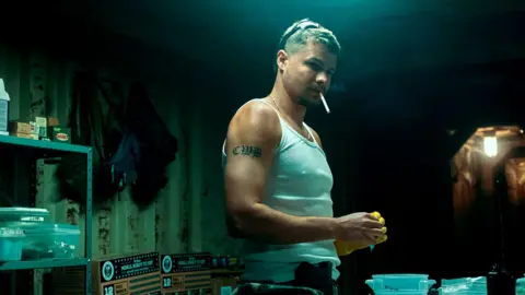 HBO Toby is wearing a white vest top, he has a tattoo on his right arm. He has chain necklace on and sunglasses on his head, he also has a cigarette in his mouth. 
