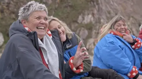 Braving the Blue - Our Lives, BBC Cymru Wales Sian smiling and laughing sat with Jo and Eva on the beach. She has her left finger in the air and is wearing a grey jacket, a orange polka dot fleece and a grey scarf. Jo is smiling whilst leaning on Sian. Eva, in the right corner, looks at the two of them with a big smile. 