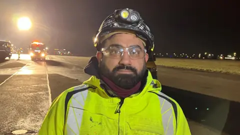 BBC/SAM READ A head and shoulders image of Suresh Patel looking at the camera. He is wearing a black helmet that has a light on the front. Behind him is a well-lit airport runway at night. Over his left shoulder construction vehicles and a large floodlight can be seen. Over his right shoulder a series of small lights can be seen in the distance.