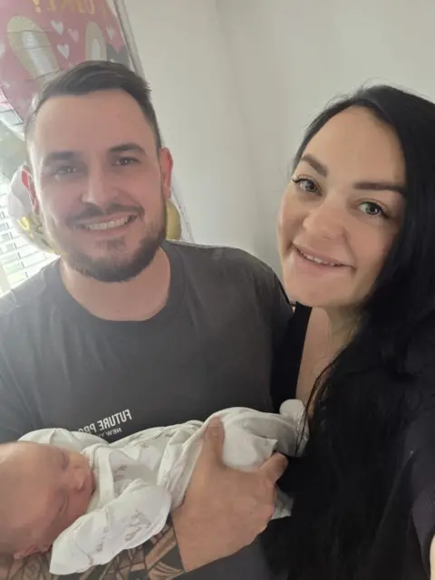 Manchester University NHS Foundation Trust Man with short dark hair and beard with a dark top with a baby wrapped in white and a woman with brown hair and a dark top. They are standing in a living room.