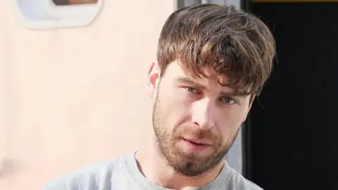 A man with medium length, messy brown hair. He is looking at the camera and wearing a grey jumper