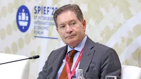Getty Images Man with brown and greying hair, wearing a grey suit with blue short and brown tie. A red lanyard is around his neck.