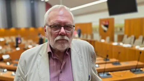 Paul Moseley/BBC Steve Morphew is wearing a light, cream linen jacket over a red and white pin-striped shirt. Behind him, there is a council chamber.