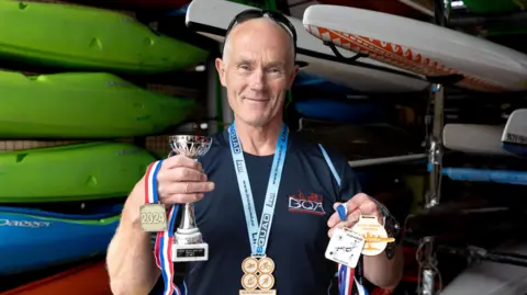 Jules Taylor standing in front of some canoes, holding three medals and a trophy. He is wearing a fourth around his beck.