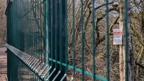 A close-up of a green fence erected at a country park. An overgrown piece of land is visible beyond it with a "private land" sign attached to a tree. A footpath can be seen on our side of the fence to the left.