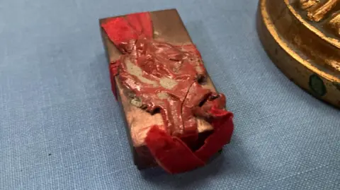 A small metal box has a red ribbon tied in a cruciform around it and the knot sealed with red wax. It lays on a blue background cloth.
