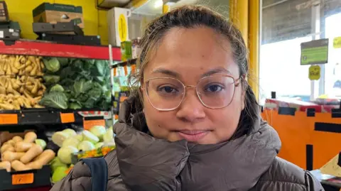 Erica Barbosa is looking directly at the camera with a neutral expression on her face. Her hair is tied back and she is wearing metallic glasses and a puffy brown coat pulled up high around the neck. There are vegetables stacked high on the shelves around her.