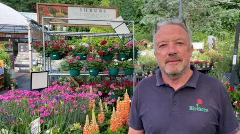 Steven Ainscough, the managing director of Birkacre Garden Centre, is photographed wearing a navy blue polo shirt. He has short, grey hair and a trimmed beard. His glasses are resting on top of his head. He stands in front of colourful plants in the garden centre.