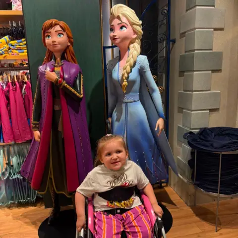Charlie Brown A young blonde girl in a black wheelchair with black straps across her chest. She is wearing a white Wizard of Oz t shirt and pink and orange striped trousers. Behind her are life-size figures of Disney pricesses Anna and Elsa.