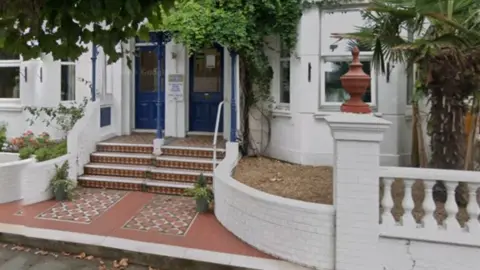 A white terraced house with two blue doors next to one another, with a palm tree on the right in the front garden